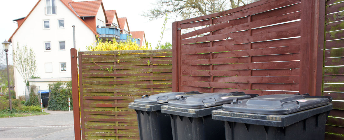 Foto: drei Restabfallbehälter stehen am Holzzaun einer Müllstandsfläche 